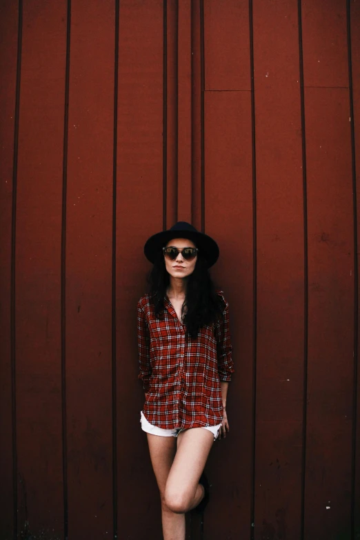 a woman standing against a wall in a red plaid shirt and black hat