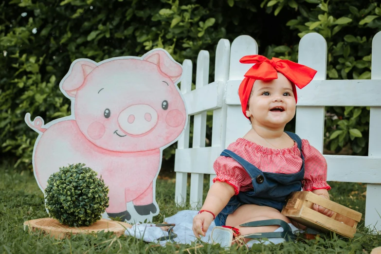 a baby girl is sitting in the grass by a fence with a little pig cutout