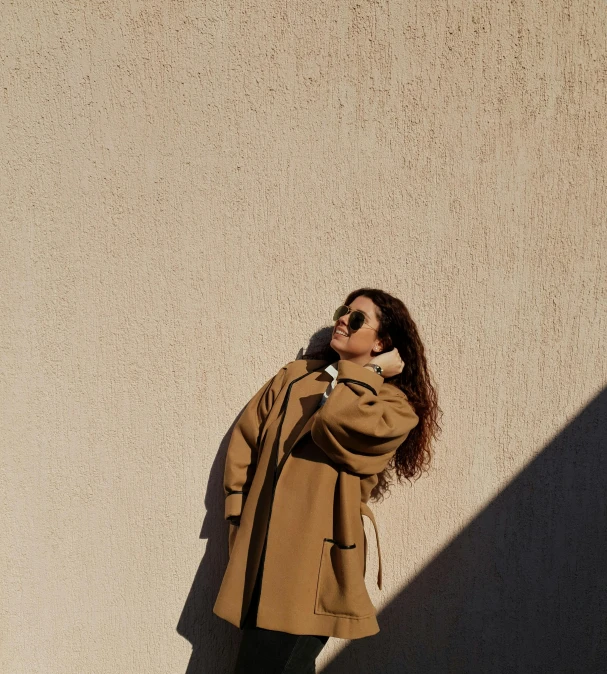 a woman in a brown coat and black boots standing against a building wall