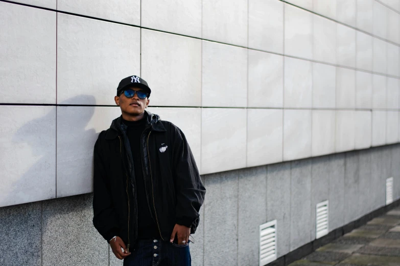 man in blue sunglasses and cap leaning against wall