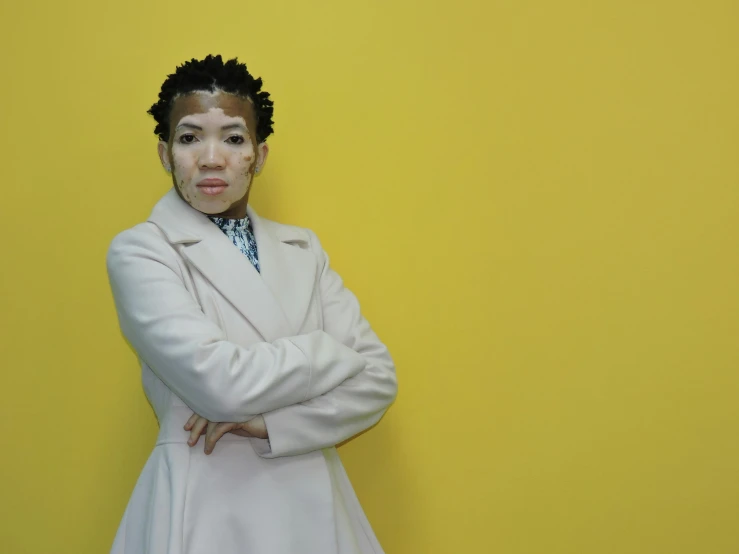 a young woman in white coat posing against a yellow wall