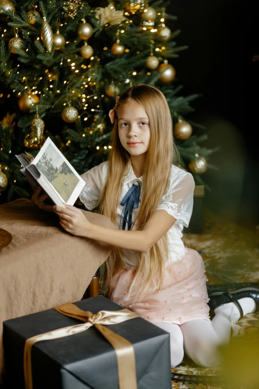 the girl is reading a book by the christmas tree