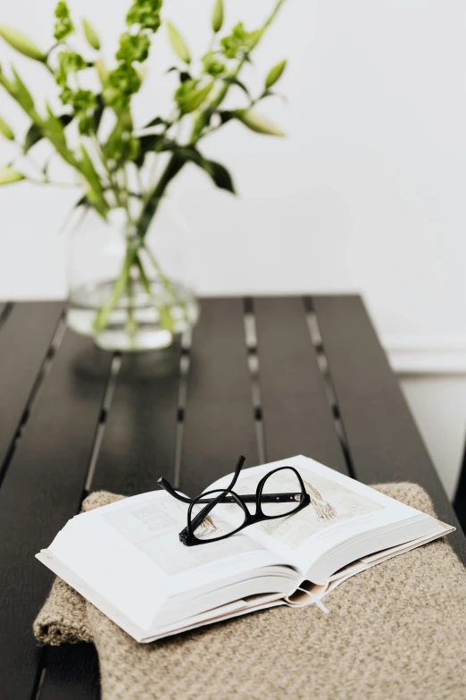 an open book with glasses on it, next to a vase of flowers