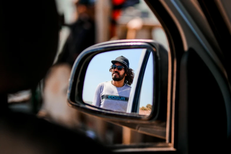 a bearded man looks out his side window as he reflects in the rear view mirror