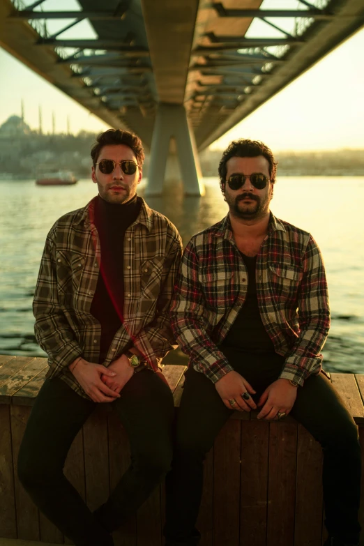 two men wearing sunglasses sitting on the dock by a bridge