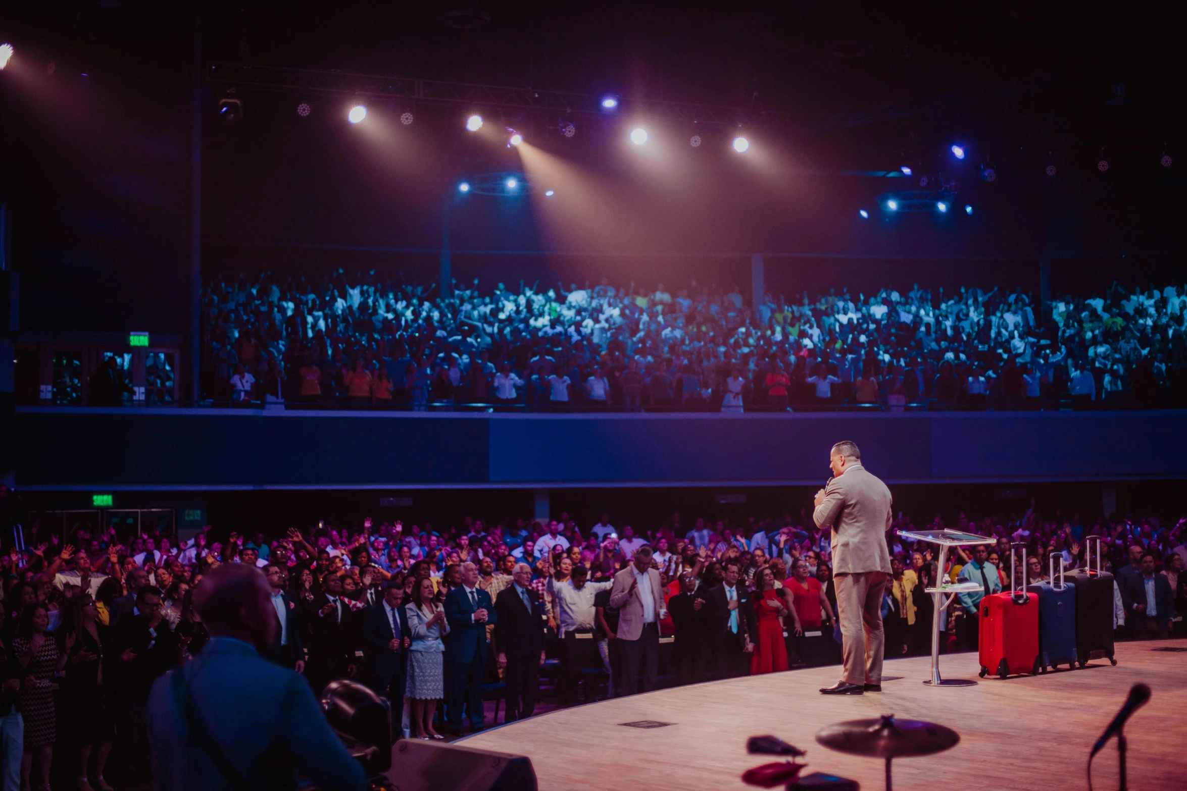 a man in a white suit and crowd behind him is speaking on a stage