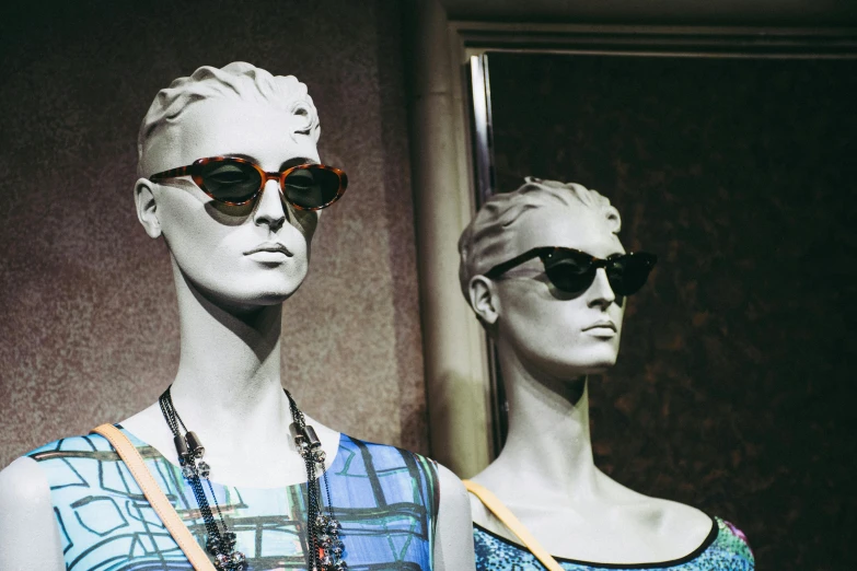 two mannequins, each wearing sunglasses and a dress, stand side by side in front of a mirror
