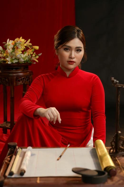 a woman in a red dress looks at the camera with pens and paper next to her