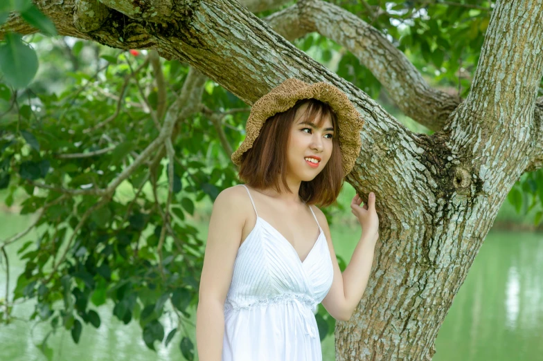 the woman wearing a straw hat stands next to a tree and looks over her shoulder