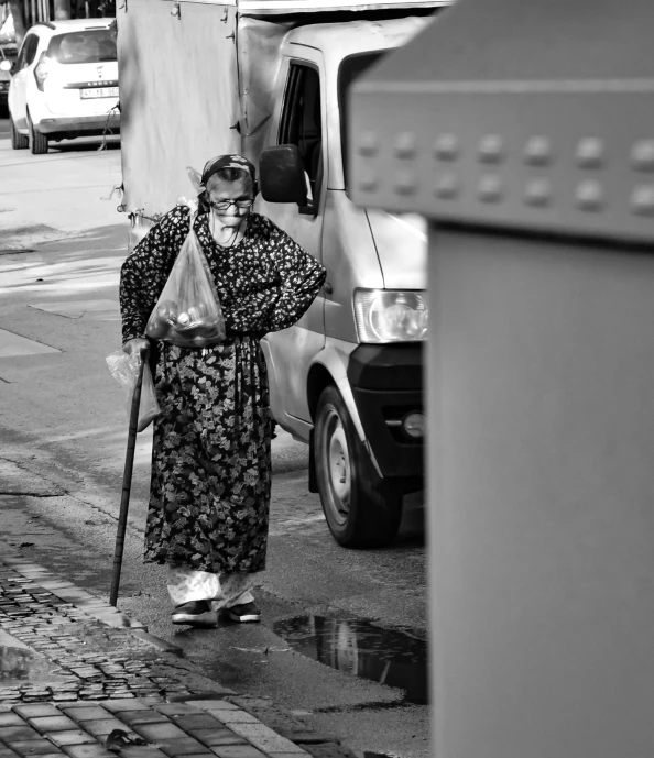 an old woman with a cane, dressed in a long gown and holding a bag is on the street