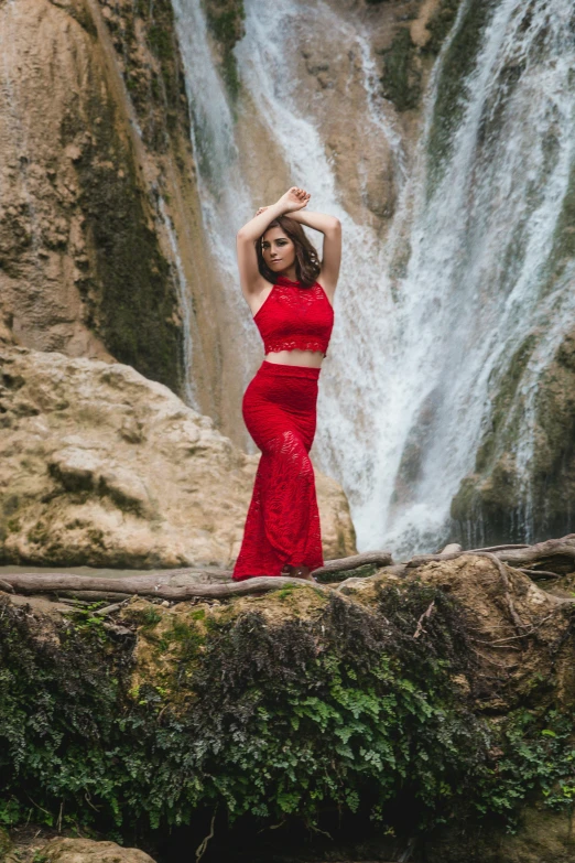 a young woman in a red dress stands near a waterfall