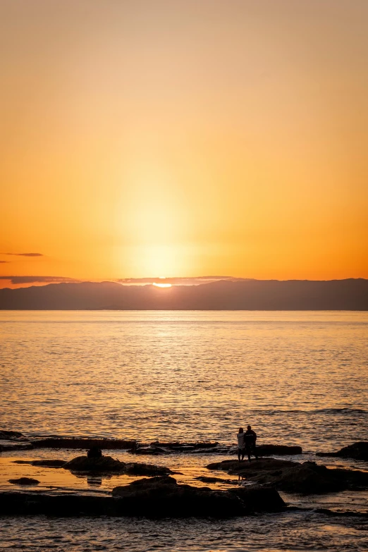 two people in the distance sitting on the edge of a rock looking at the sunset