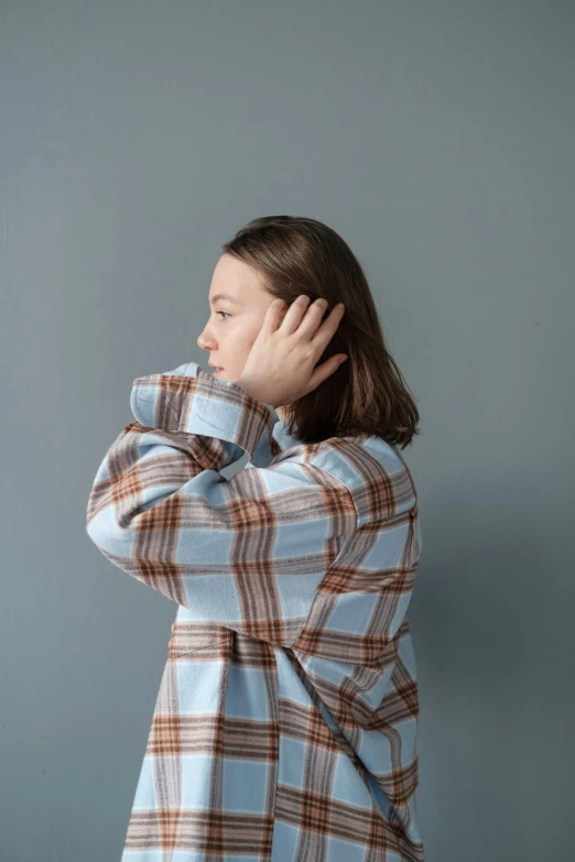 the woman stands in front of a gray wall, her hands on her head and looking off to the right