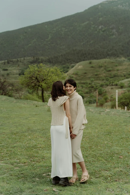 two women stand back to back in a field with hills and trees behind them