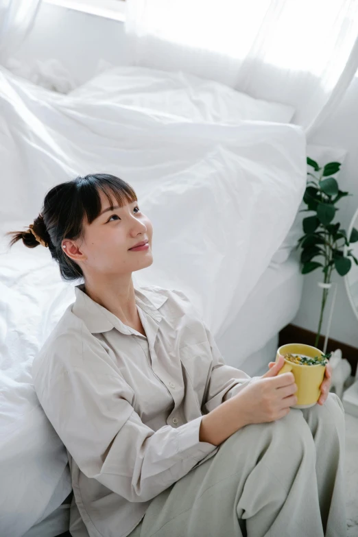 woman wearing beige sitting in the corner of bed holding a cup