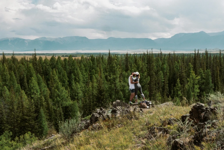 two people walking across a grass covered hillside near pine forest