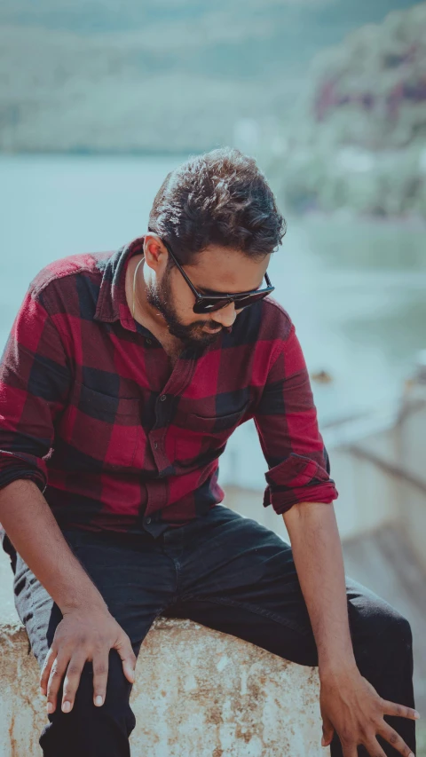 a man wearing sunglasses sitting on a rock by a lake