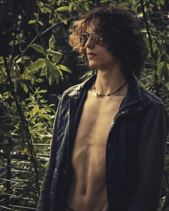 a shirtless man wearing jeans and a leather jacket stands in front of trees