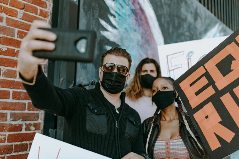 a man in a black jacket and mask is holding up his cellphone while two woman stand next to him