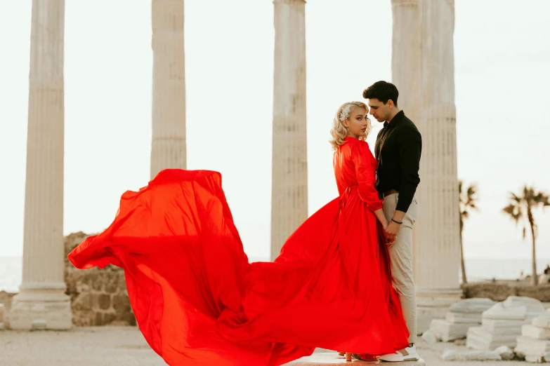 a couple holds their red dress up in the air, as they walk through some pillars