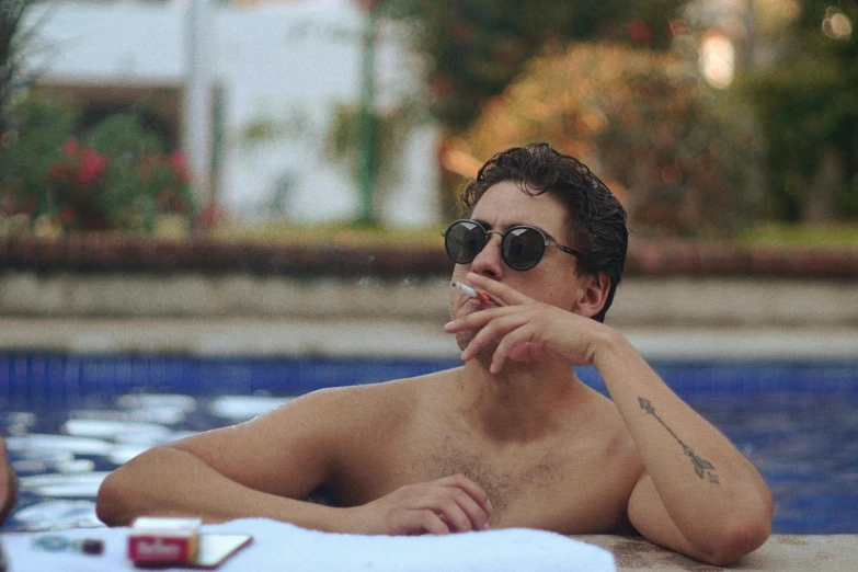 a shirtless man with sunglasses sits next to a swimming pool