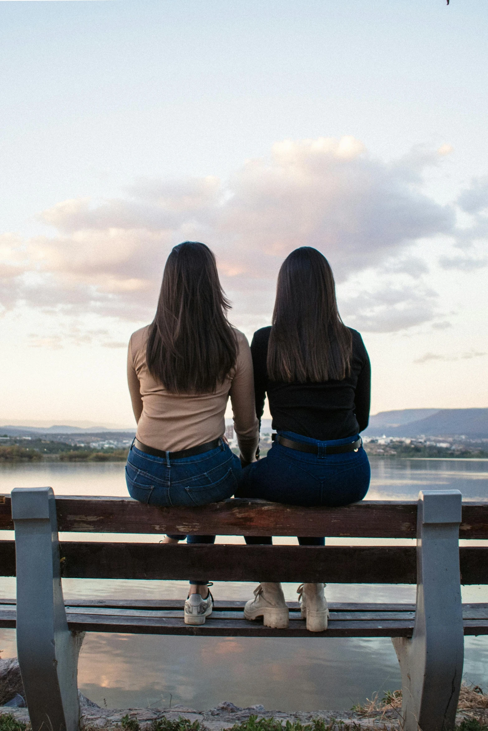 two girls sitting on top of a bench looking out over water