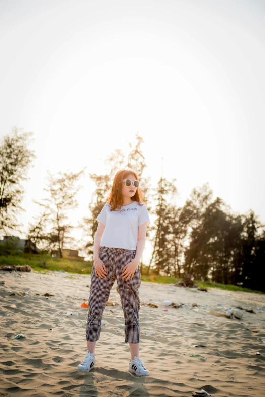 a woman wearing red headband, plaid pants and white t - shirt stands on the beach as the sun sets