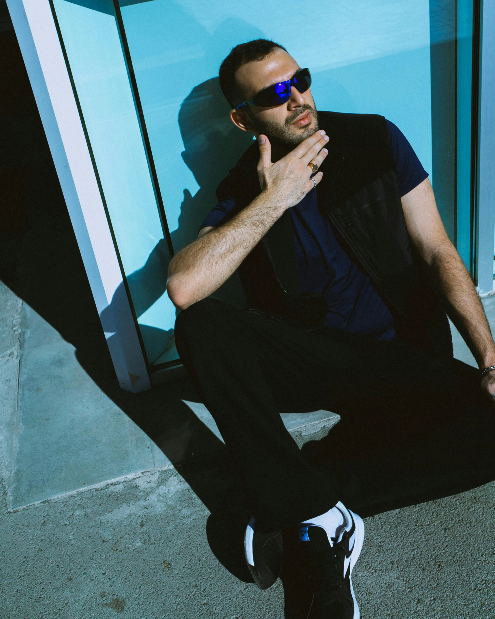 the man in sunglasses sits on the pavement in front of a blue glass wall