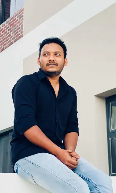 an indian man sitting on a ledge wearing blue jeans and a black shirt