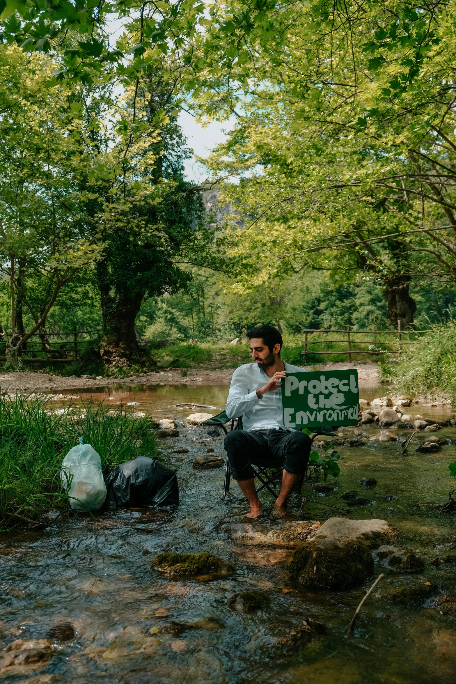 a man sits on a bench in the middle of a creek holding an eco sign