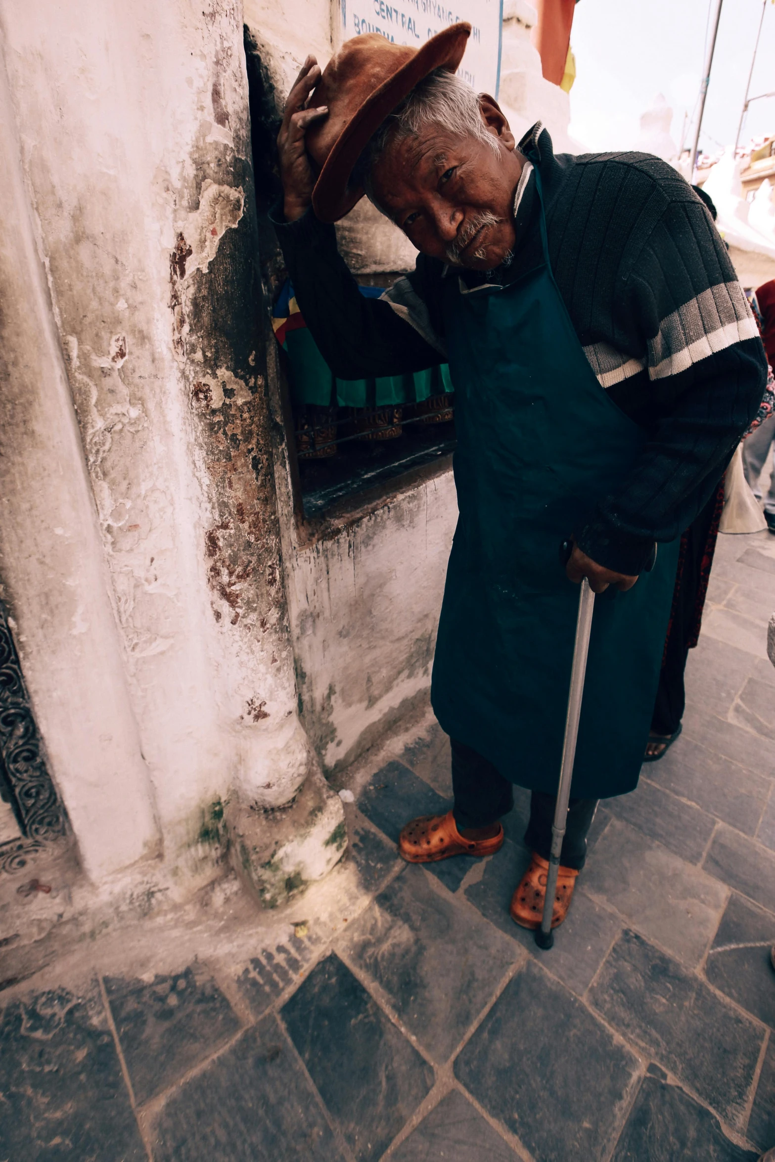 an old man leaning against a wall with a stick and a hat on his head