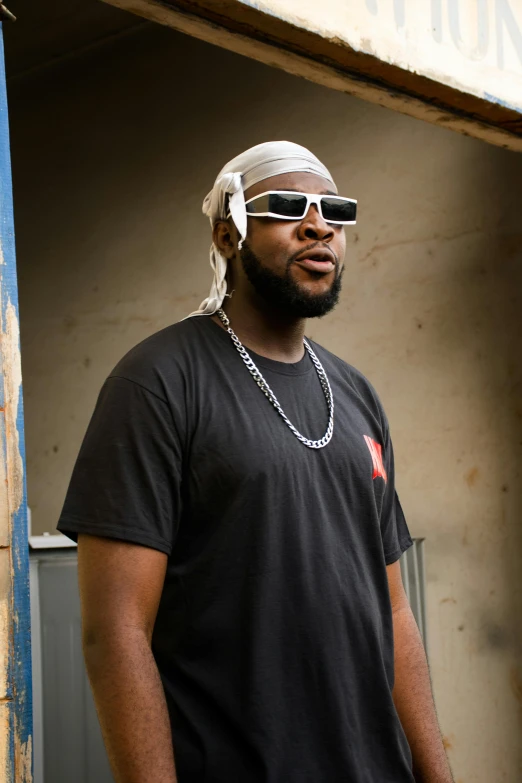 a man standing by a building wearing sunglasses and a bandanna on