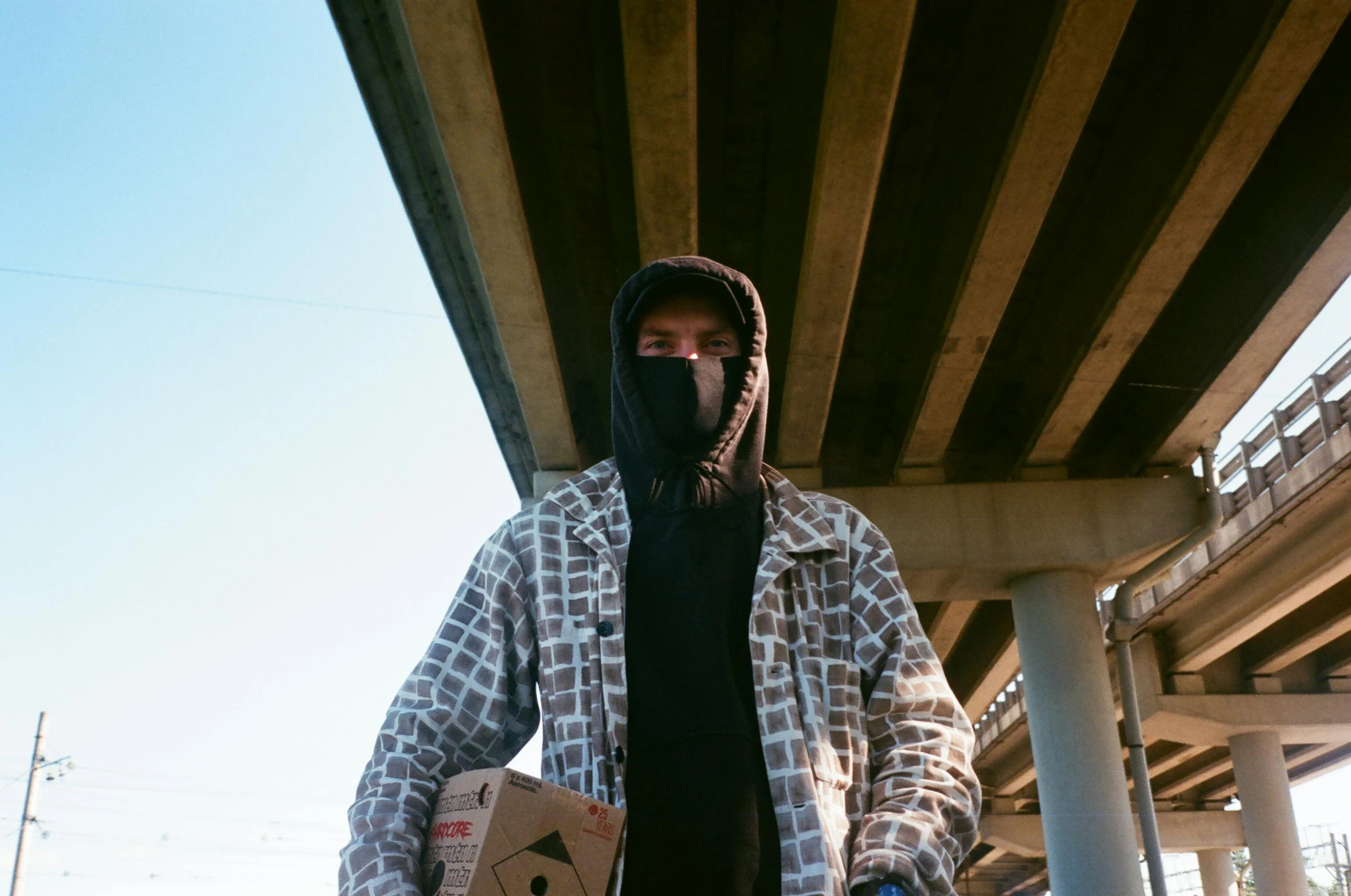 a person wearing a hoodie holds a skateboard under a bridge