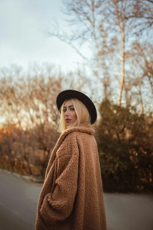 a blonde woman with a wide brimmed hat and an oversized cardigan is standing in a treed road