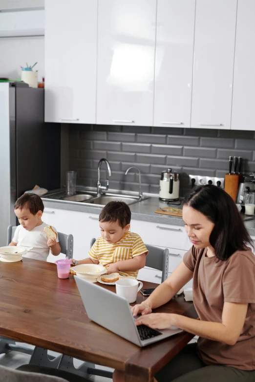 a woman sitting at a kitchen table with two children on laptops