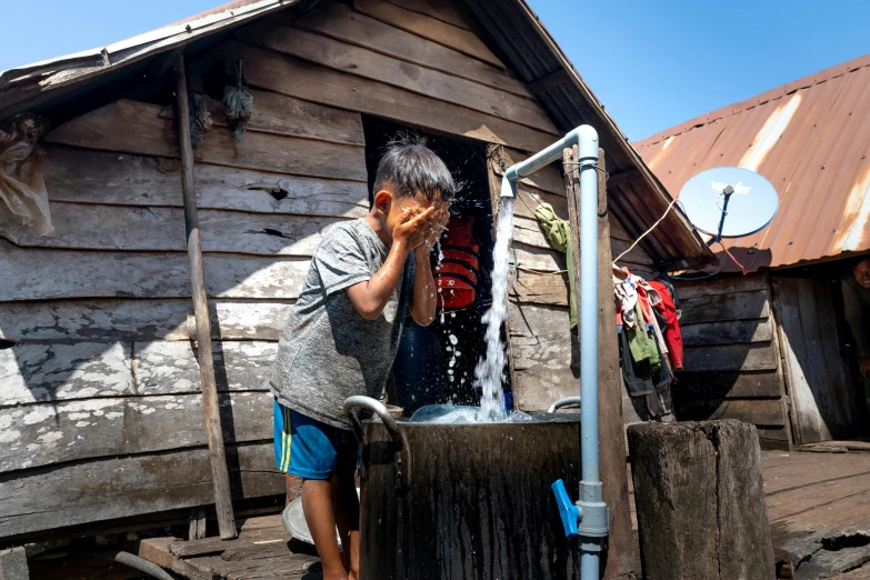 a little boy drinking out of the spigot in front of a shack