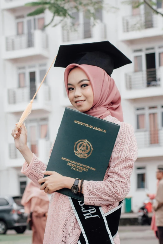 a woman in a hijab holding up a green, yellow and black graduation book