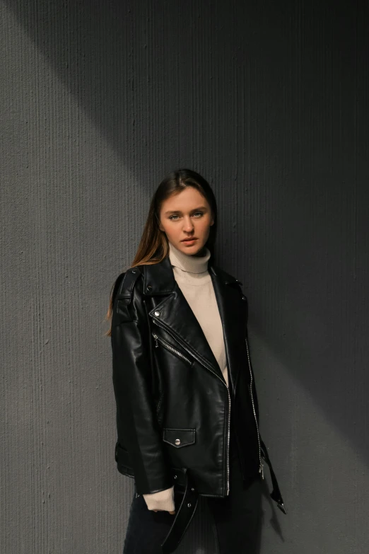 a woman posing against a grey wall in a black jacket