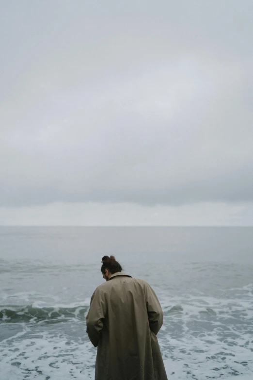 a woman in a trench coat stands at the edge of a beach