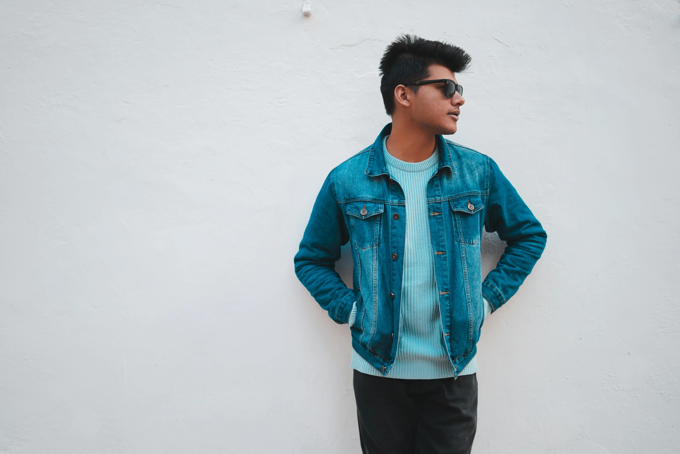 man with eye glasses standing against white wall wearing blue shirt and jeans