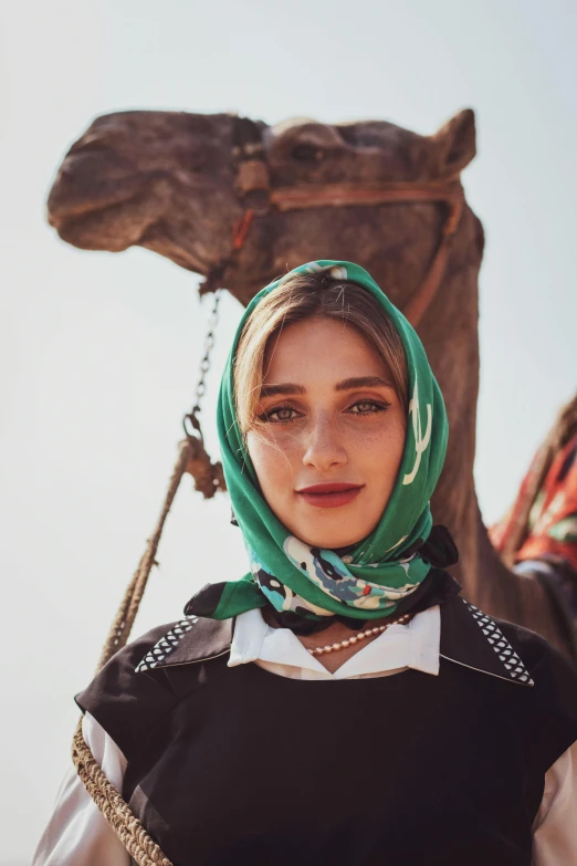 a woman in a black top and green scarf stands in front of a camel