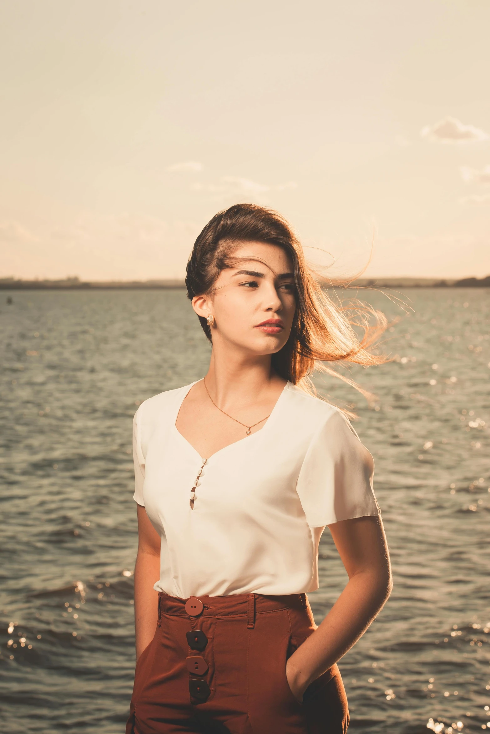 a beautiful young lady in a white top and brown pants stands in front of the water