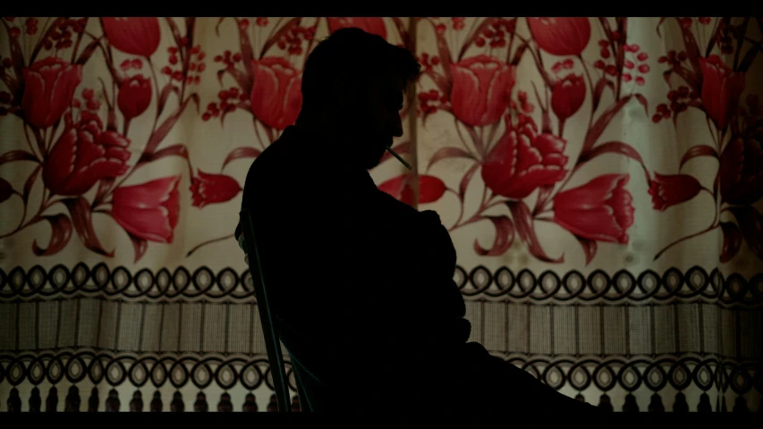 a man is sitting in a chair in front of a wall with red floral design