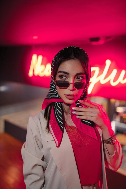 a woman in sunglasses and a scarf standing under a neon sign