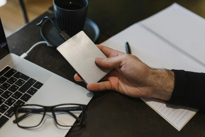 man's hand holding notebook with white cover while using laptop