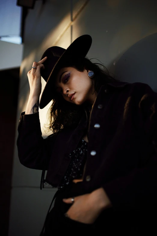 a beautiful young woman in a black jacket and hat leaning against a wall