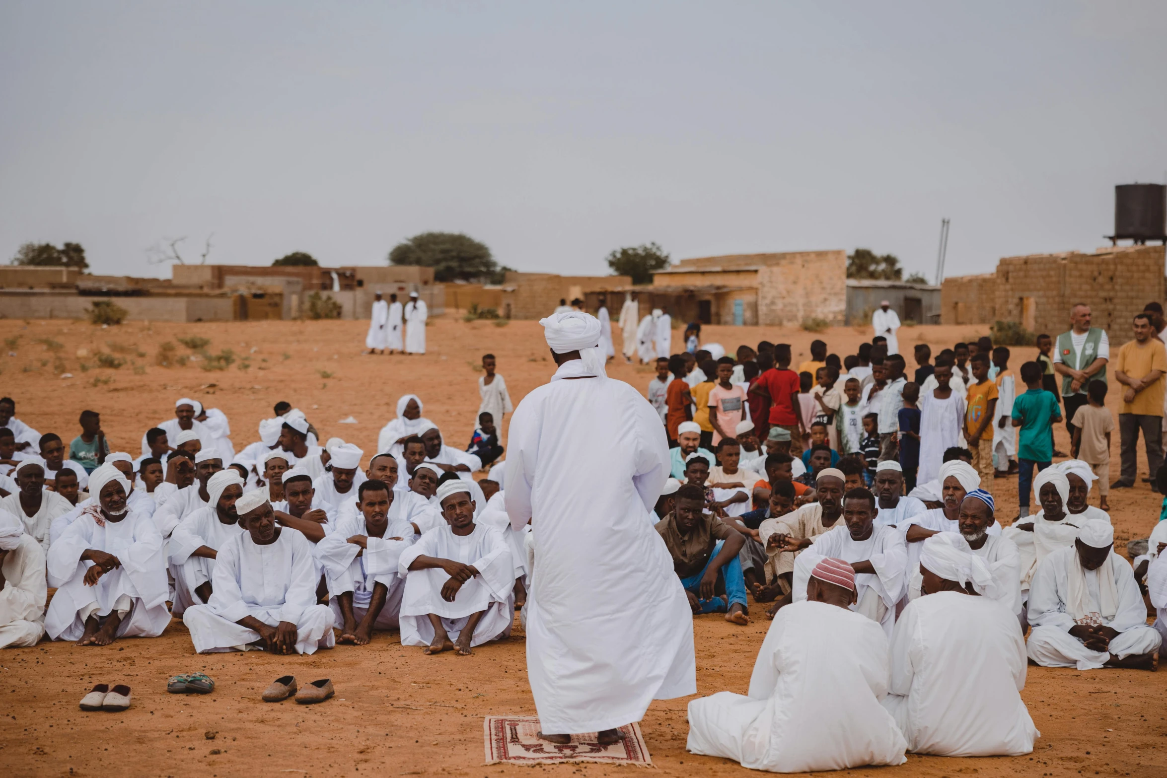 people and many dressed in white sit on the sand as they stand by a building