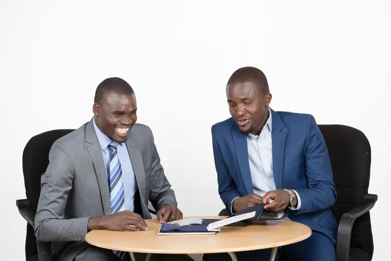 two african men sitting at a table in suits looking at a magazine