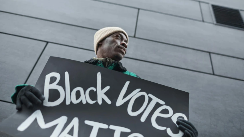 a man holding up a black voting sign with words written on it