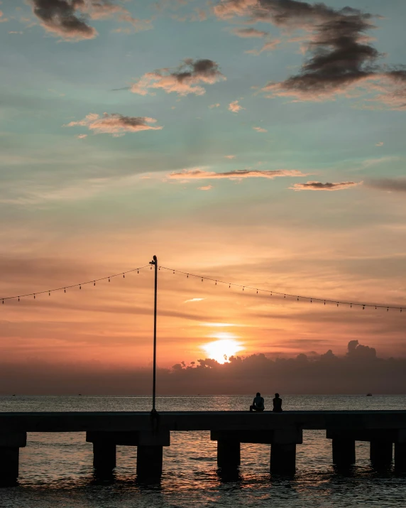 a couple of people are sitting on a pier watching the sunset